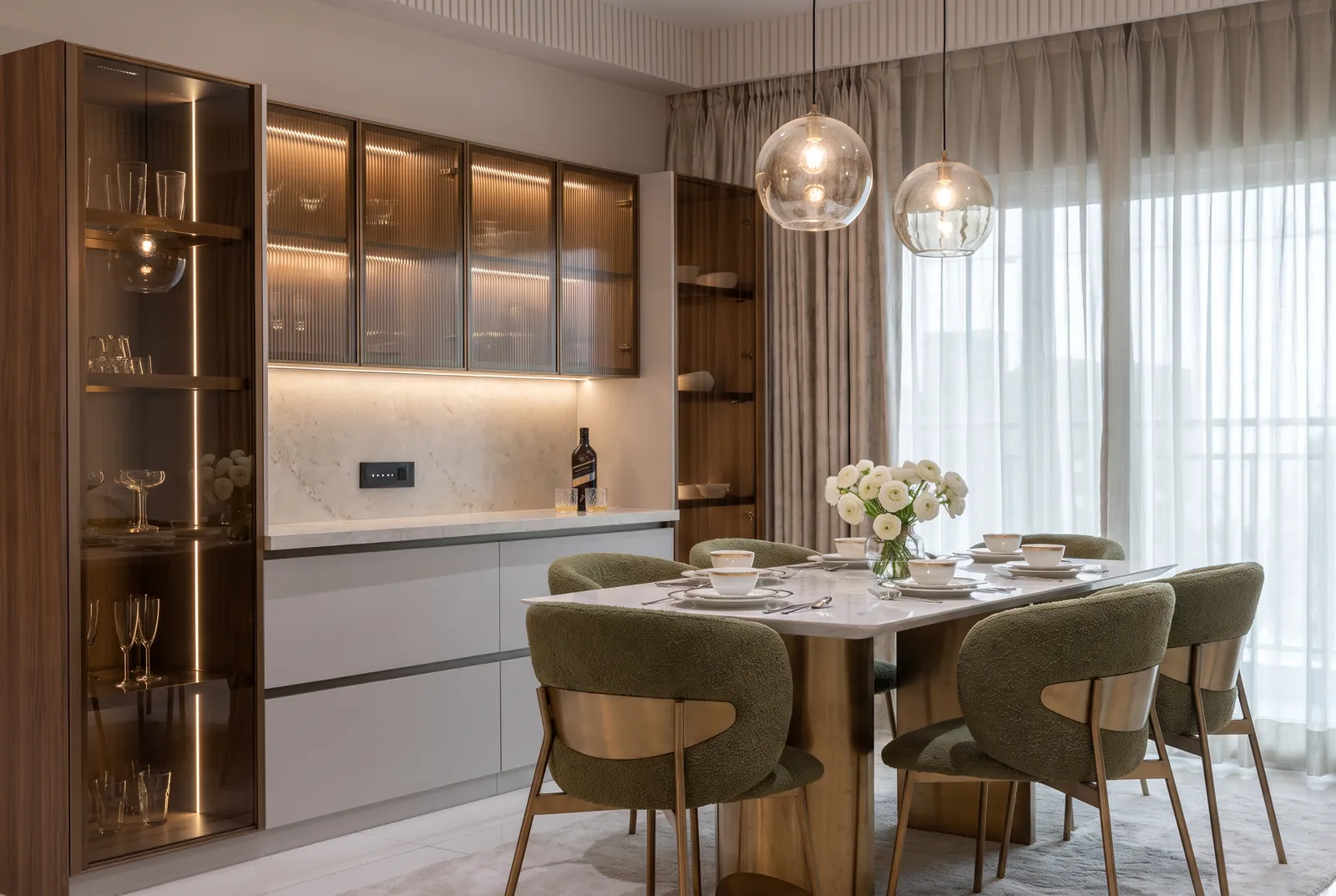 A dining table with olive boucle chairs in front of a bar wall of bronze fluted glass and walnut display cabinets.